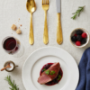 Cubertería con baño de oro total sobre una mesa con mantel blanco y un plato con un guiso de carne.